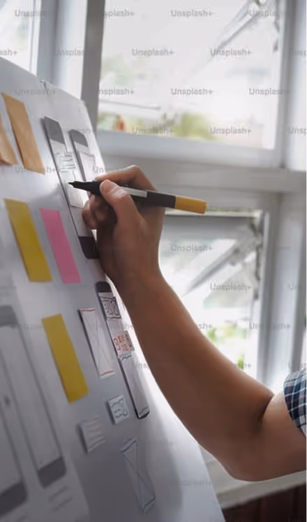 Person holding a black and yellow marker, drawing or writing on a whiteboard with sticky notes and sketches near a window.