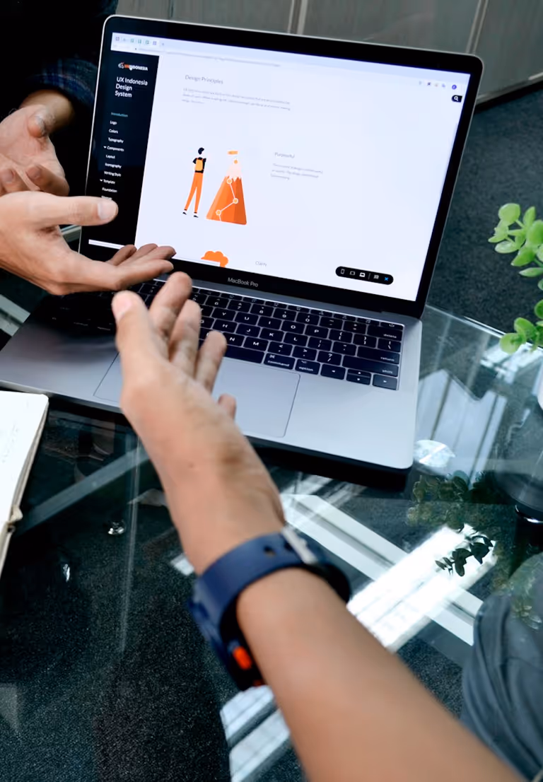 Two people discussing UX Indonesia Design System displayed on a MacBook Pro screen on a glass table.