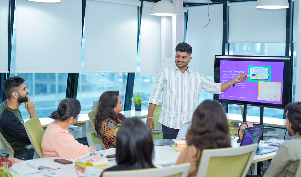 A young man in a striped shirt presenting concept sketches on a screen to a group of six colleagues seated around a conference table in a modern office.