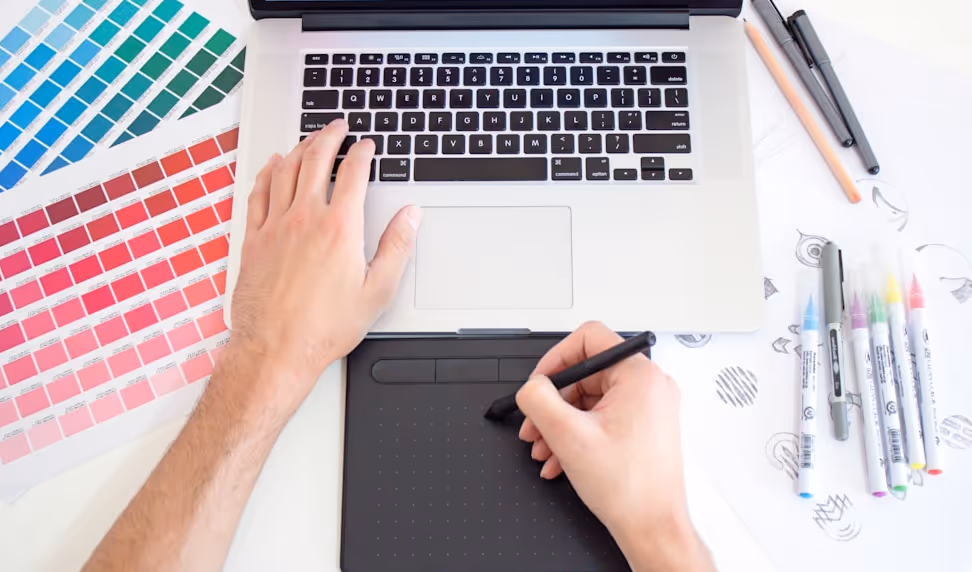 Person using a graphic tablet and pen in front of a laptop keyboard surrounded by color swatches and markers on a white desk.