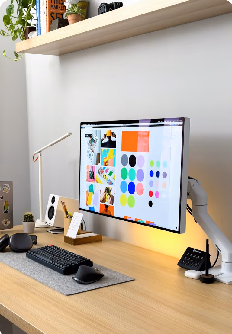 Modern desk setup with a widescreen monitor displaying colorful design elements, black keyboard and mouse on gray mat, headphones, speakers, and a wooden shelf with books and plants above.