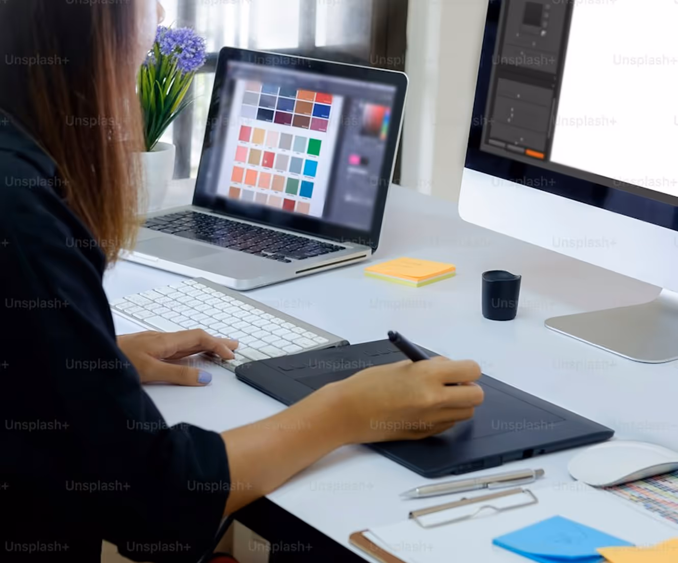 Person using a digital drawing tablet with a pen, working at a desk with a laptop displaying a color palette and a desktop monitor.