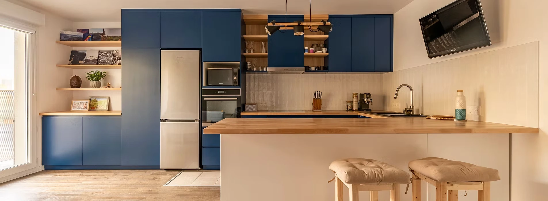 Modern kitchen with blue cabinets, wooden countertop, stainless steel fridge, and stools.