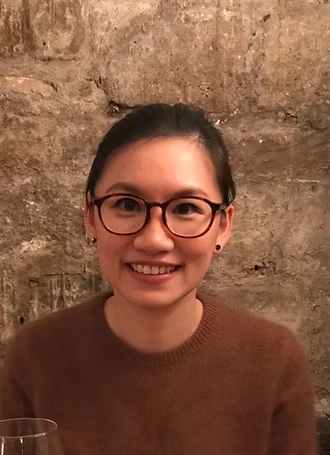 Smiling woman with glasses wearing a brown sweater against a textured stone wall.