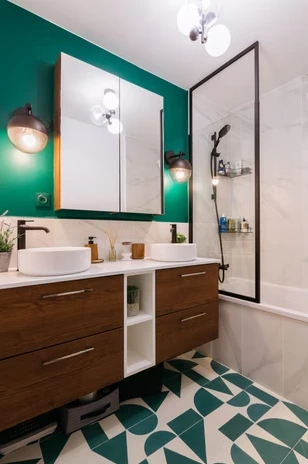 Modern bathroom with wooden vanity, twin round sinks, green wall, patterned floor, and glass shower screen.