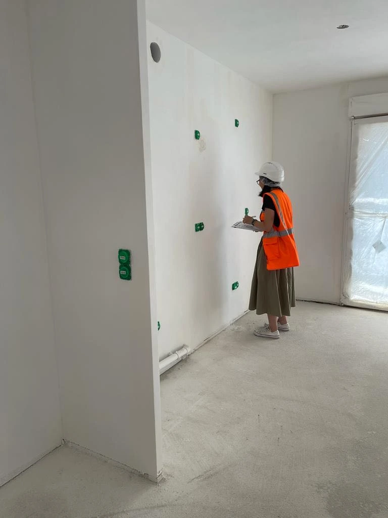 Person in a white helmet and orange safety vest inspecting white wall in empty room under construction.