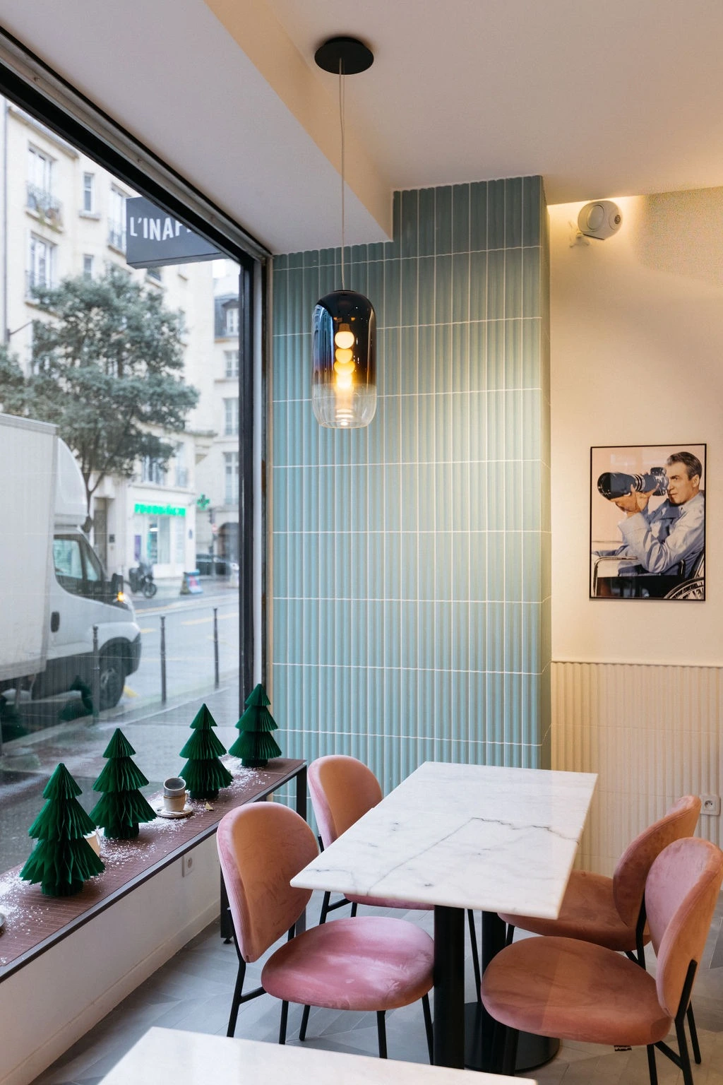 Cozy cafe corner with a marble table, pink chairs, teal tiled wall, hanging lamp, and small green trees by a window.