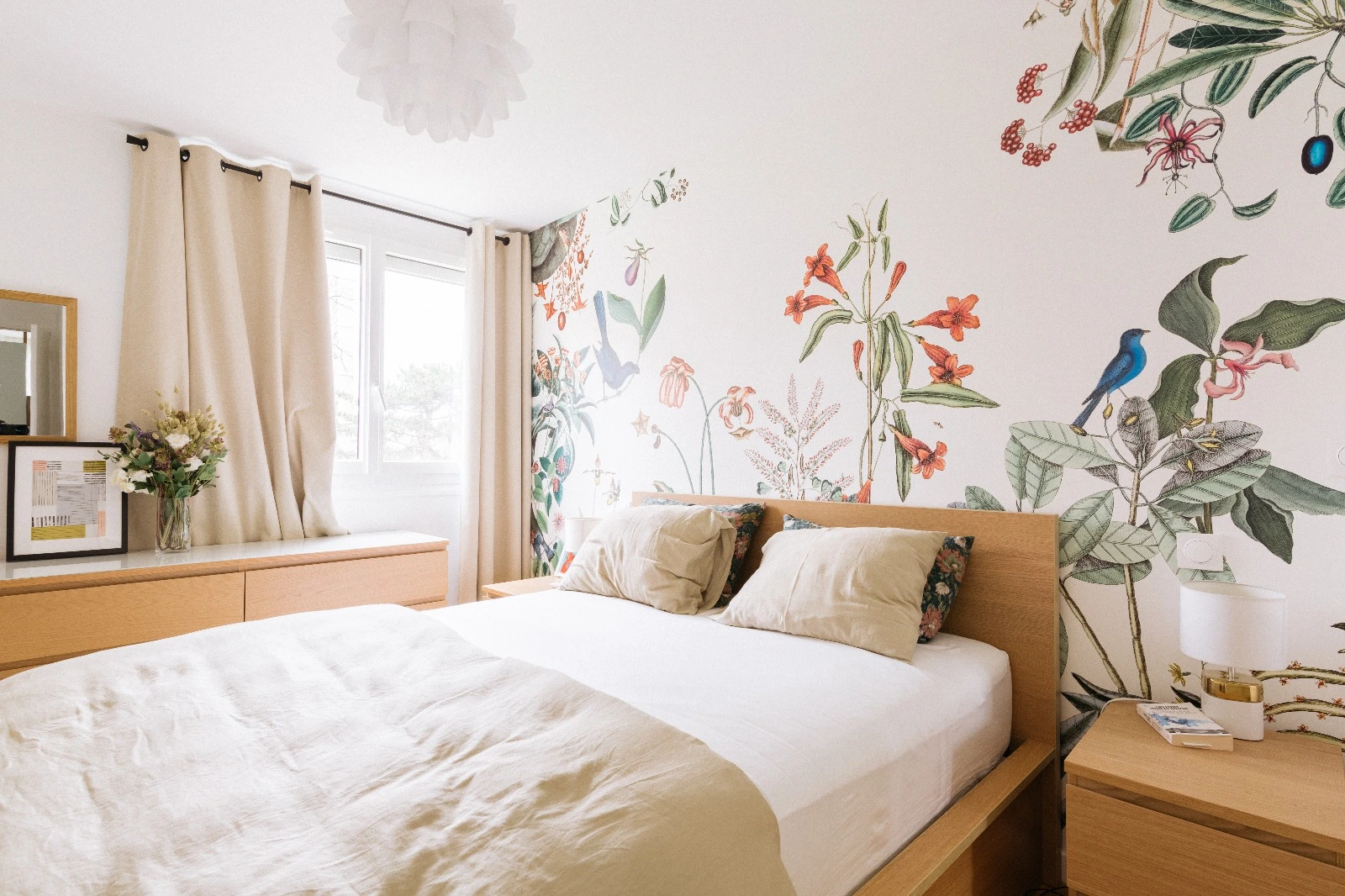Bright bedroom with wooden bed, floral pillows, botanical wall art, light curtains, and bedside lamps.