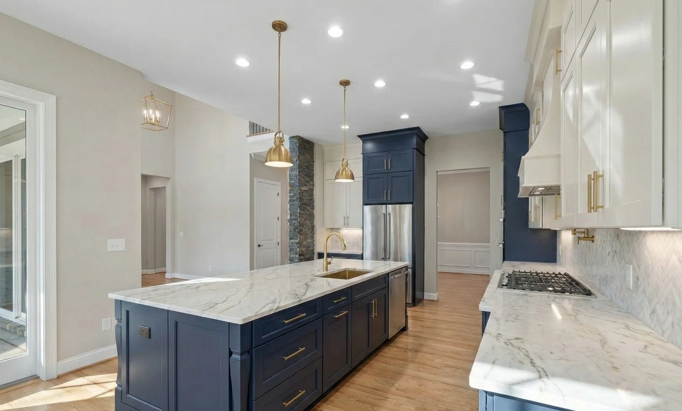 Modern kitchen with beige cabinetry, marble countertops and backsplash, stainless steel appliances, and a central island with a gold faucet.