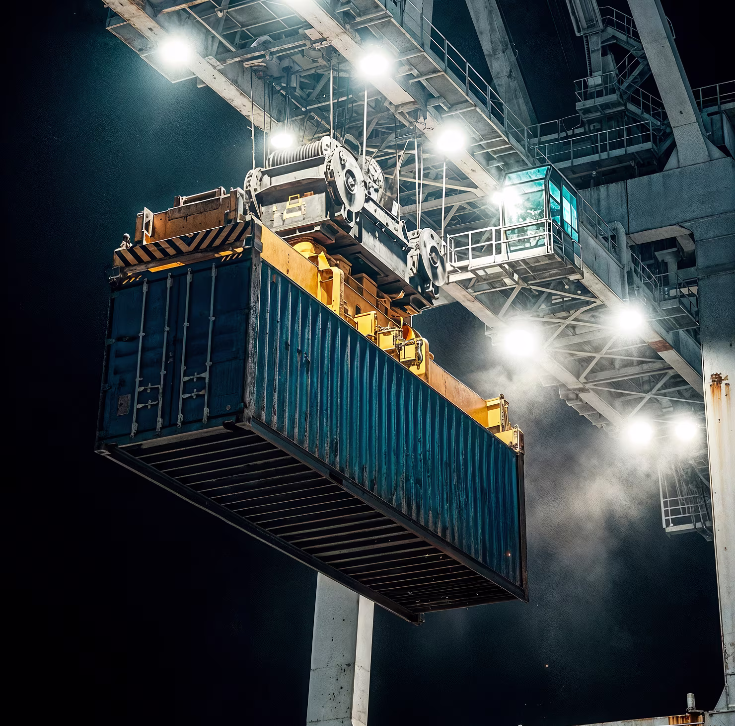 Large blue shipping container being lifted by a crane at night with bright industrial lights illuminating the scene.