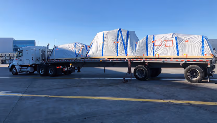 Flatbed truck parked outdoors carrying three large white objects covered with tarps secured by blue straps.