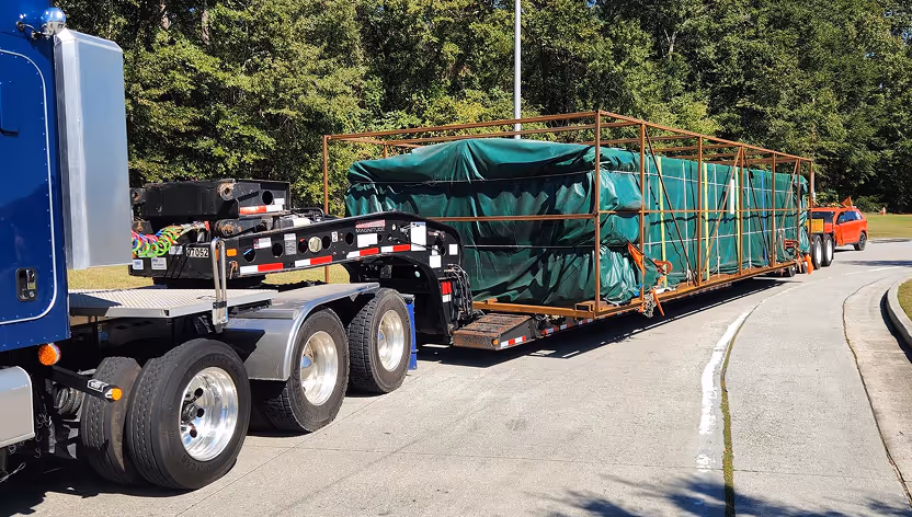 Blue semi-truck pulling a long flatbed trailer carrying large green covered cargo secured with orange straps on a paved road with trees in the background.