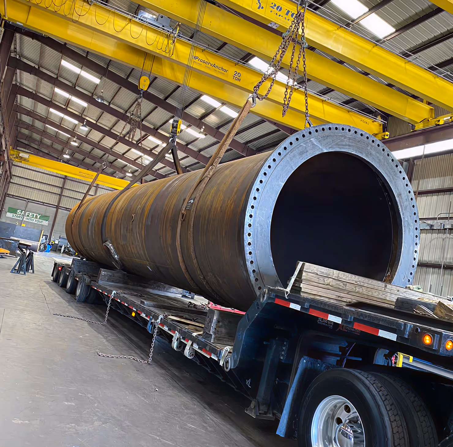 Large industrial cylindrical metal pipe being lifted by yellow overhead cranes inside a warehouse onto a flatbed trailer.