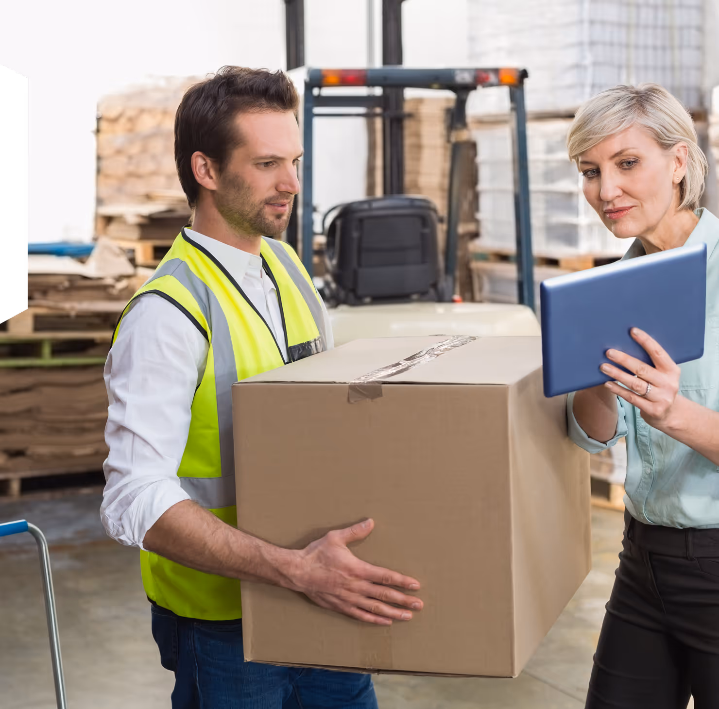 Warehouse worker in a yellow safety vest holding a large cardboard box while a woman checks details on a tablet.