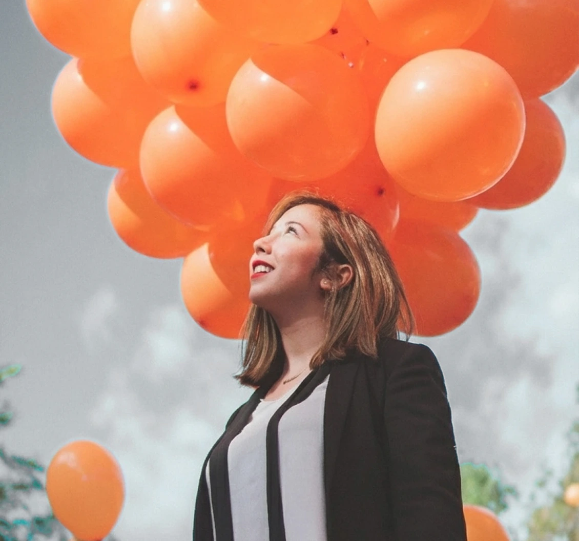 Woman looking upward while holding a cluster of orange balloons outdoors against a cloudy sky.