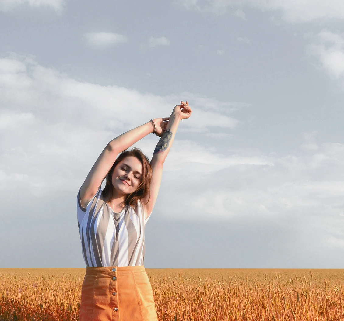 Woman standing in a golden field with arms raised, eyes closed, under an open sky, expressing calm and freedom.