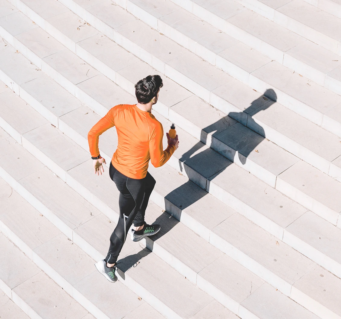Person in an orange top running up wide concrete steps, holding a water bottle, with a long shadow cast in sunlight.