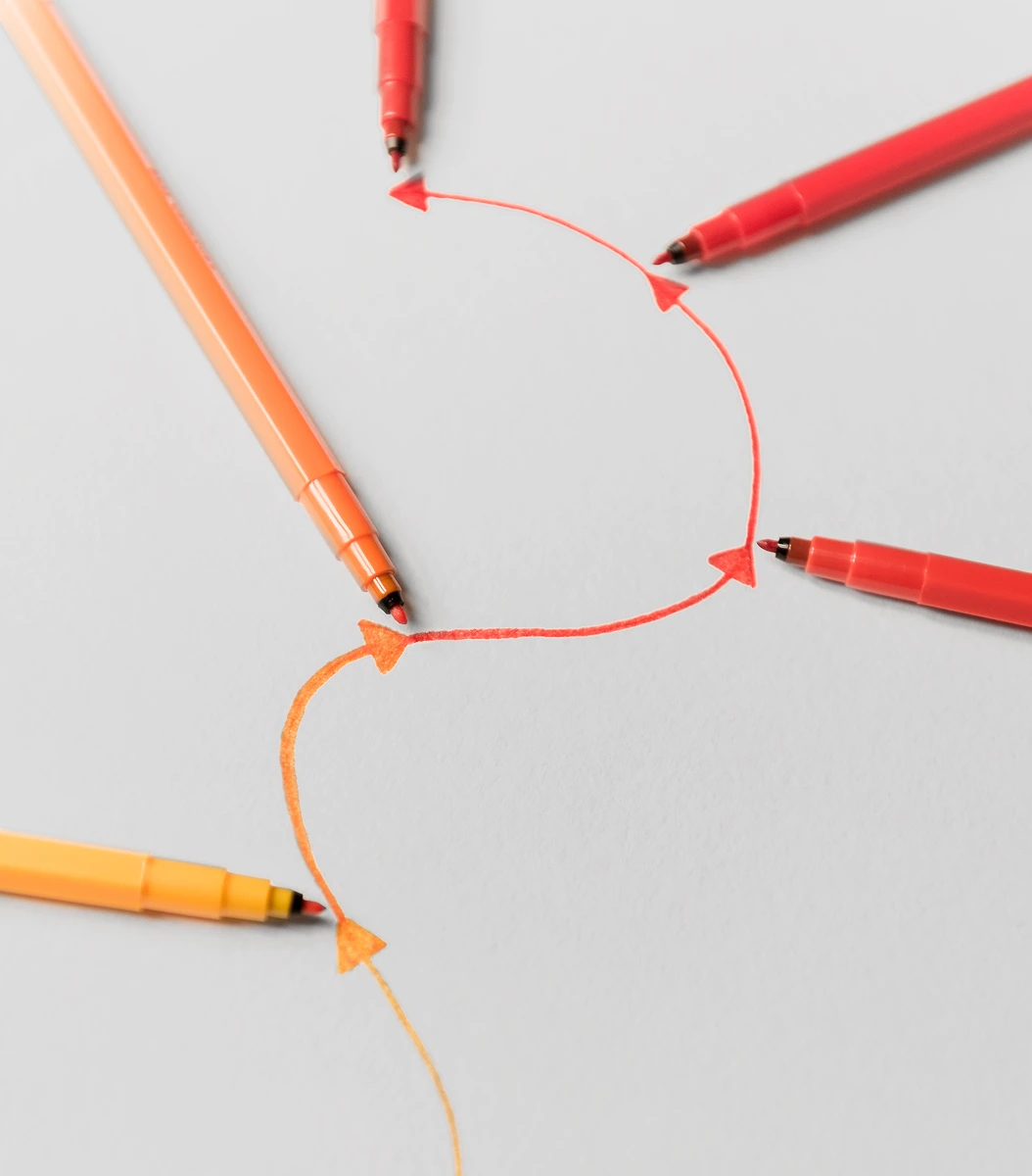 Colored markers drawing connected curved arrows on white paper, symbolizing process, flow, or direction.
