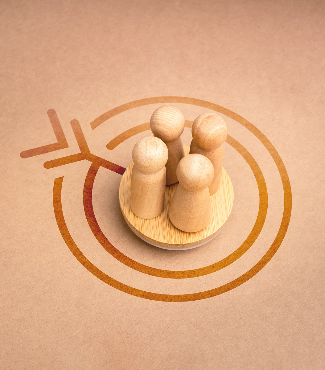 Four small wooden peg figures standing close together on a round wooden base, centered within concentric circular lines resembling a target or signal, on a warm beige background symbolizing a group, community, or focused collaboration.