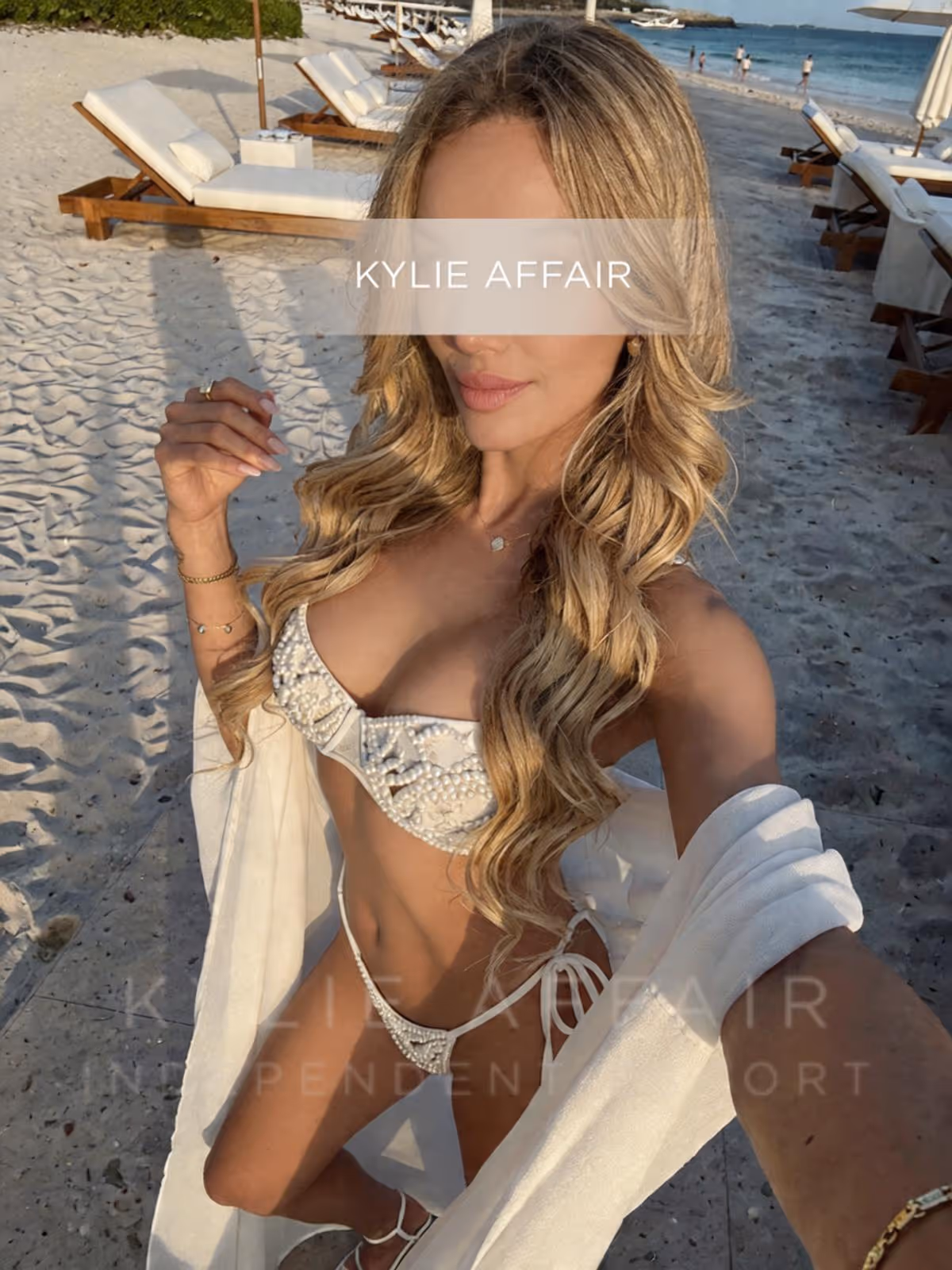 Kylie Affair in white embellished bikini and cover-up taking a selfie on a sandy beach with lounge chairs.