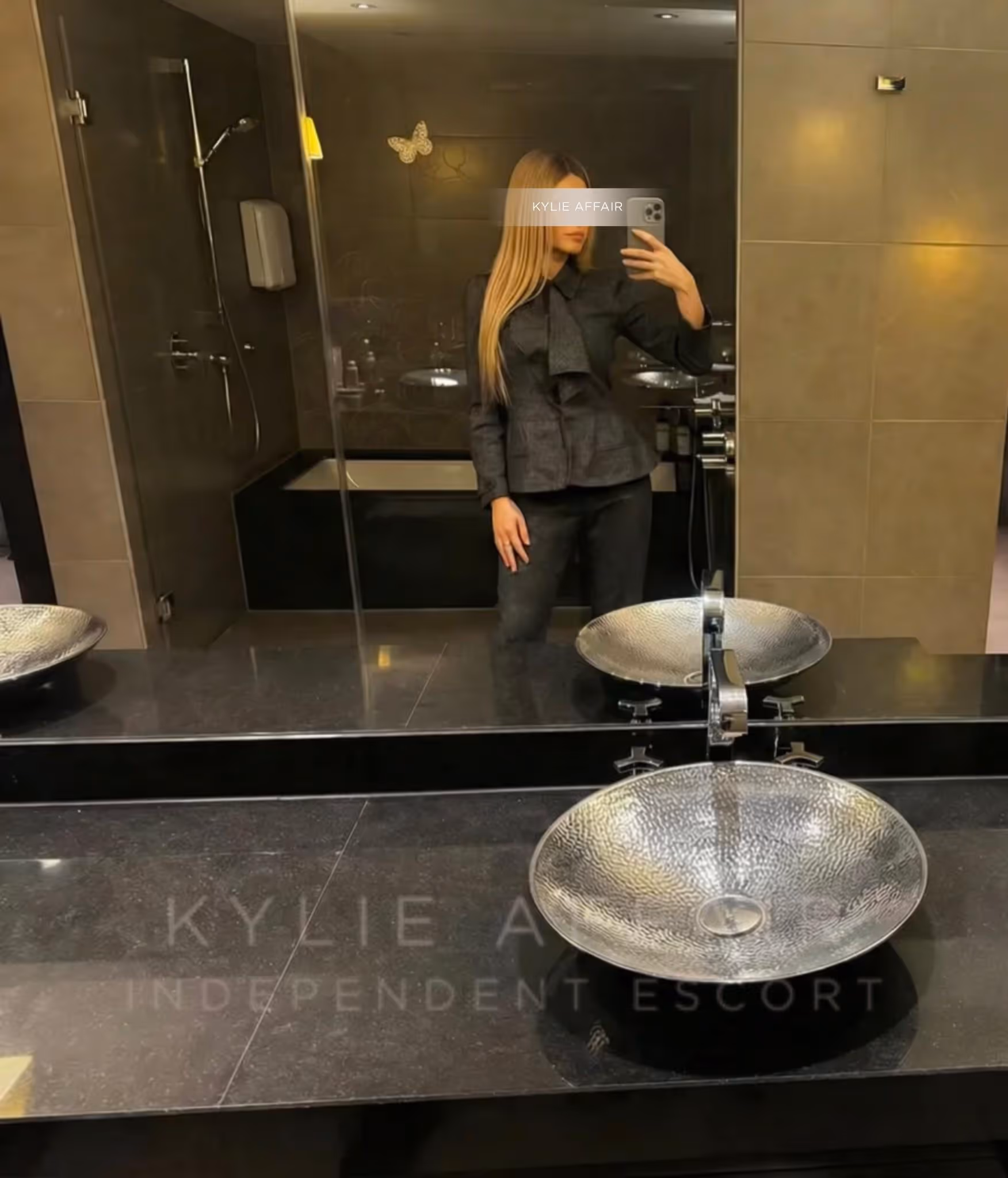 Woman with long blonde hair in black outfit taking a mirror selfie in a modern bathroom.