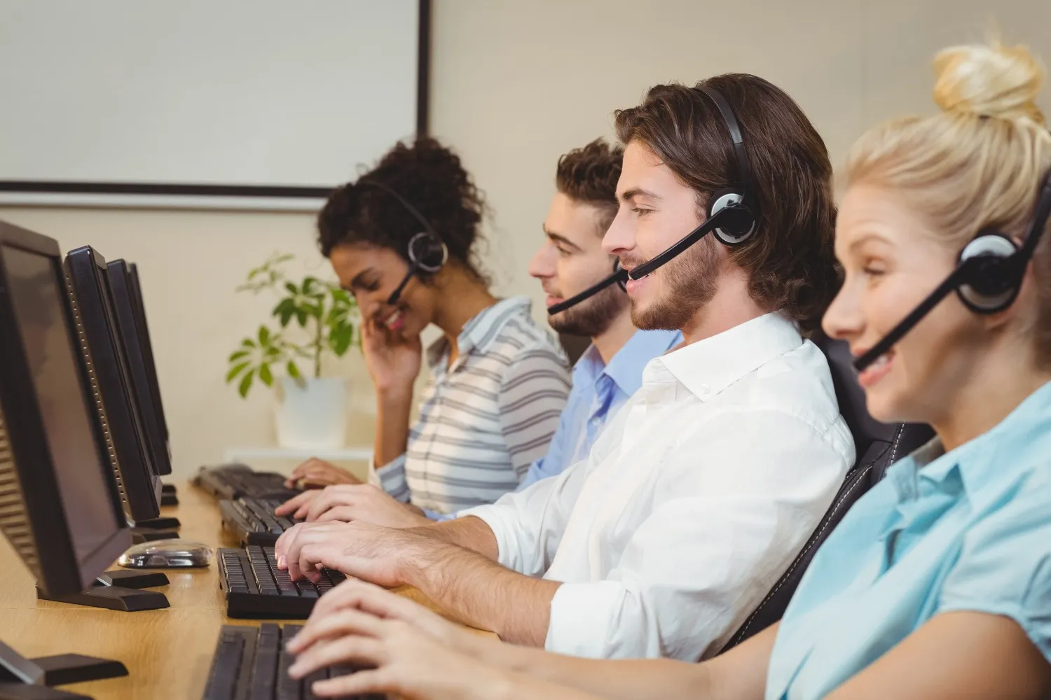 Four call center employees wearing headsets and typing on computers in a row.
