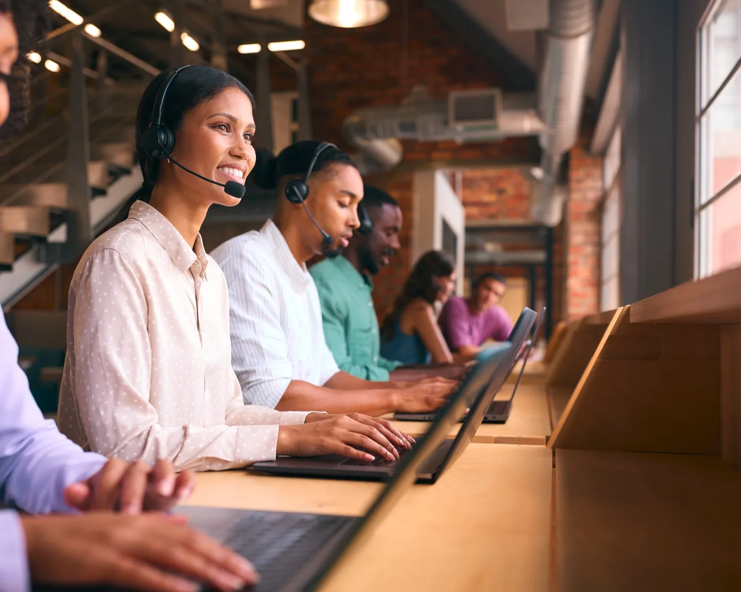 Smiling female call center agent wearing a headset and working on a laptop alongside diverse colleagues.