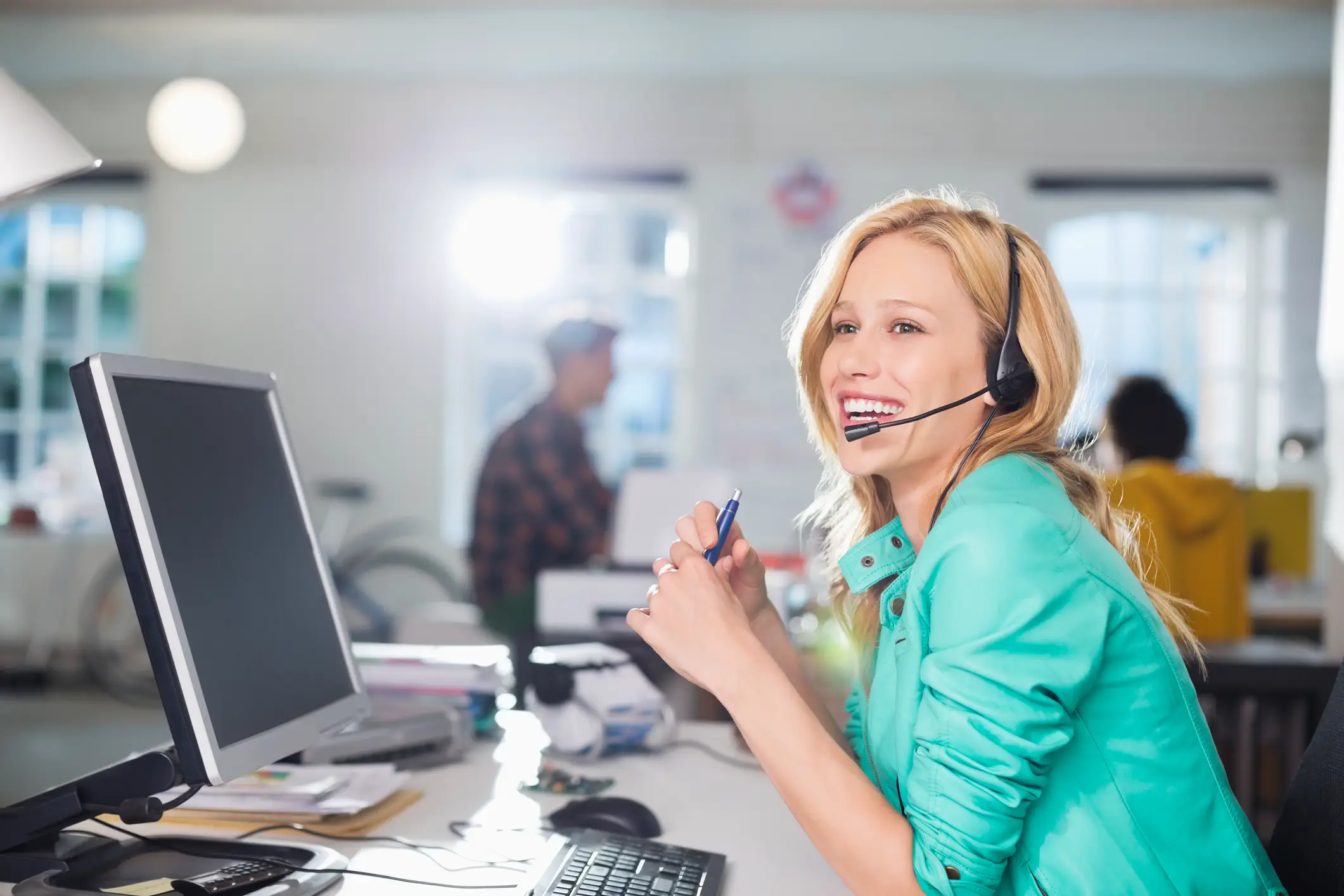 Cheerful blonde woman in a turquoise jacket and headset holding a pen in a bright, modern office.