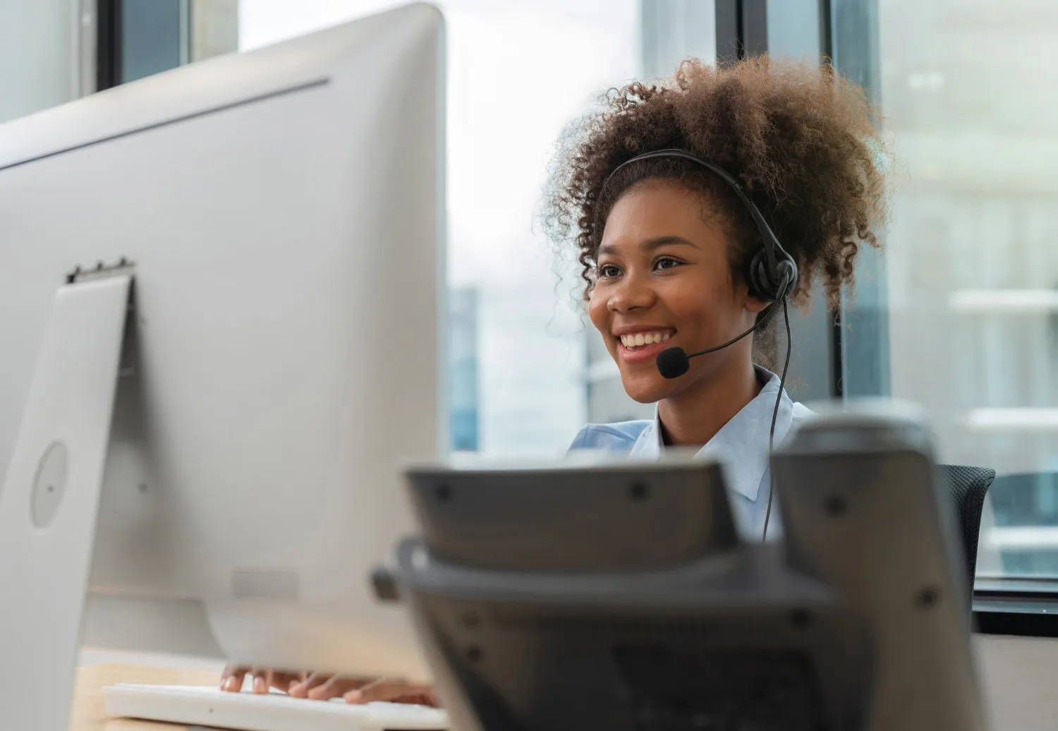  A young woman with curly hair, wearing a headset, smiles warmly while looking at a large computer monitor in an office setting. She appears to be a customer service or call center agent. The background shows a bright window with a blurred cityscape. A desk phone or similar device is visible, blurred, in the foreground.