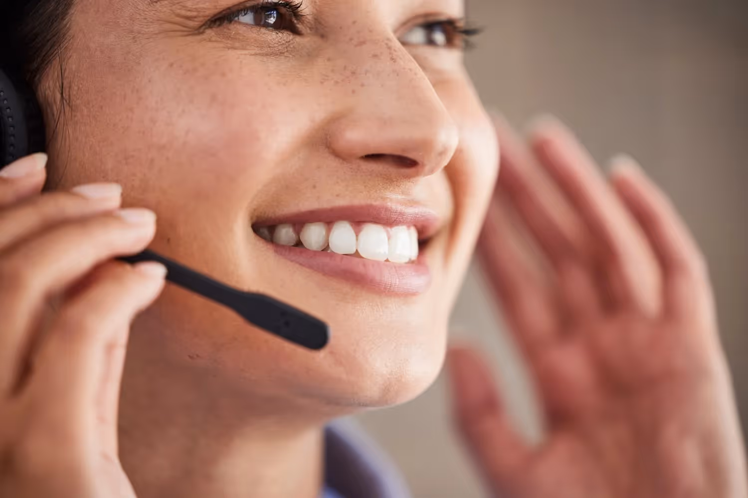  A close-up photograph focuses on the mouth, nose, and cheek of a smiling woman with freckles, who is wearing a headset with the microphone positioned near her mouth. Her hand is slightly raised near her ear, and her bright white teeth are visible as she smiles genuinely.