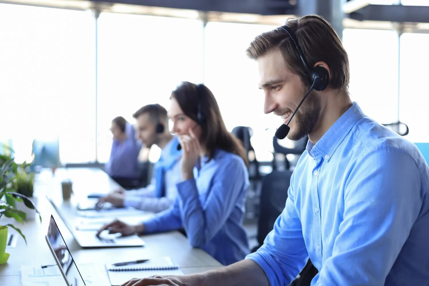 A smiling male call center agent with a beard, wearing a headset and a light-blue shirt, is working at a computer in an open office. Behind him, other agents are also on calls.