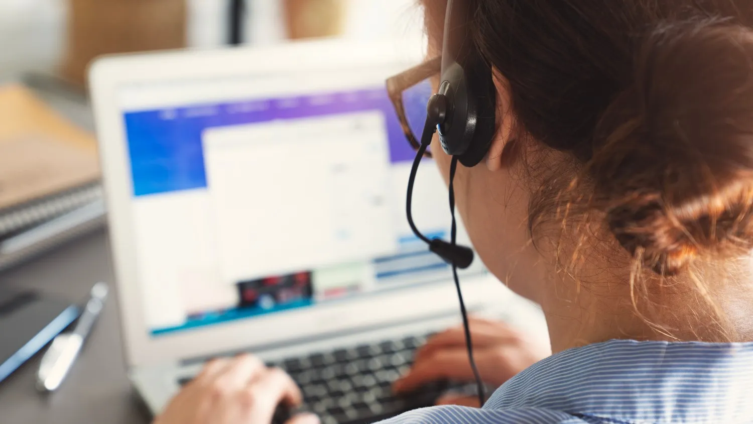  A close-up shot from behind shows a person, presumably a customer service agent, wearing a headset and spectacles, typing on a laptop computer. The laptop screen displays a web page with a pop-up window. The person's hair is tied back in a bun, and they are wearing a light-colored shirt.