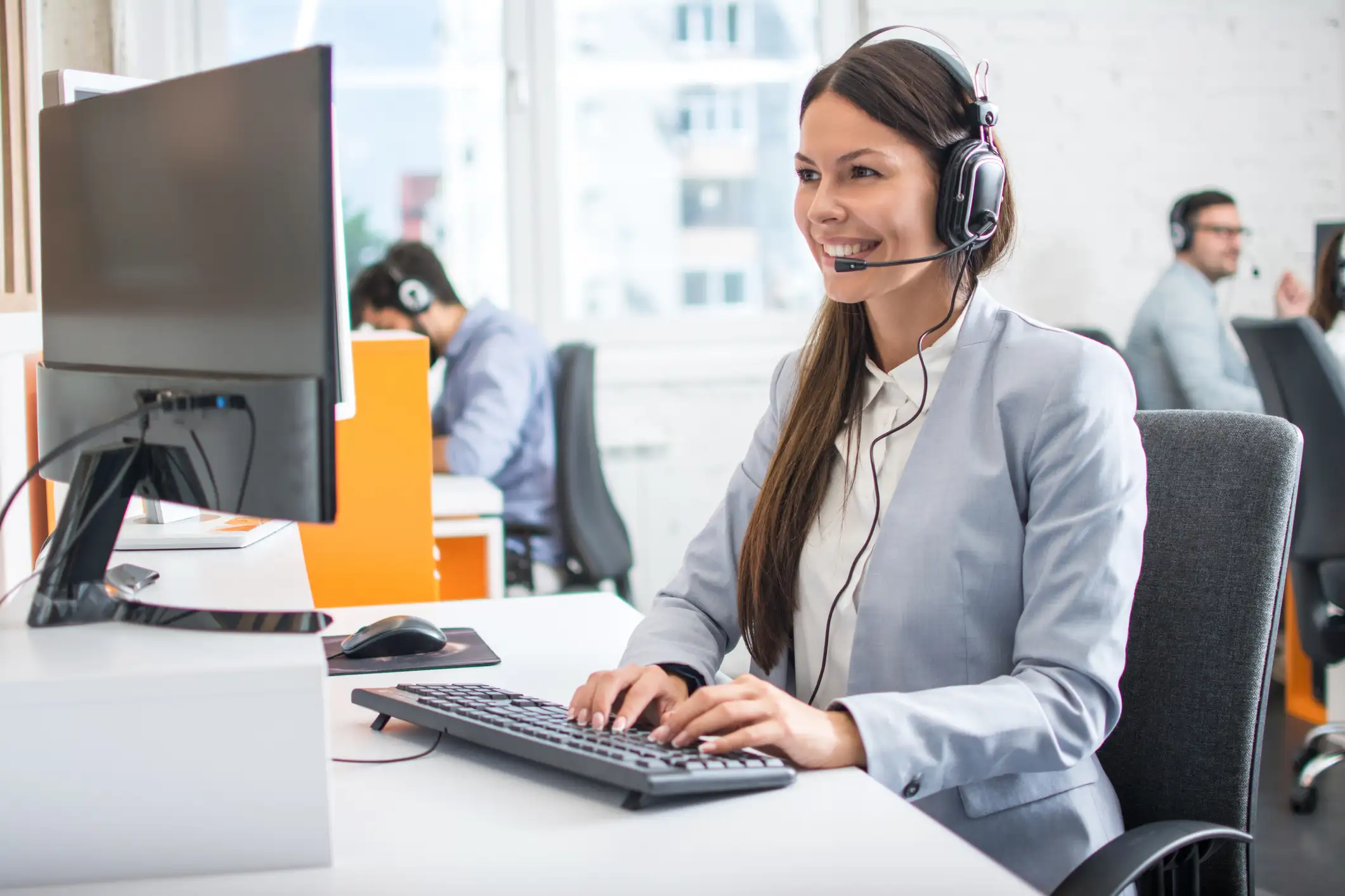 Professional woman with long brown hair and a headset typing on a keyboard in a bright call center.