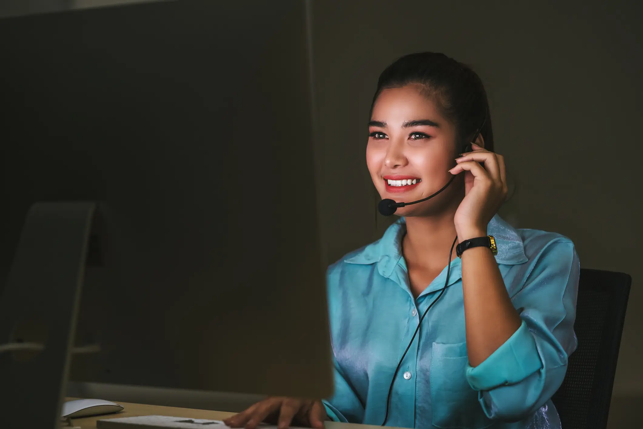 Woman in a blue shirt smiling and working late on a customer service call in a dark office.