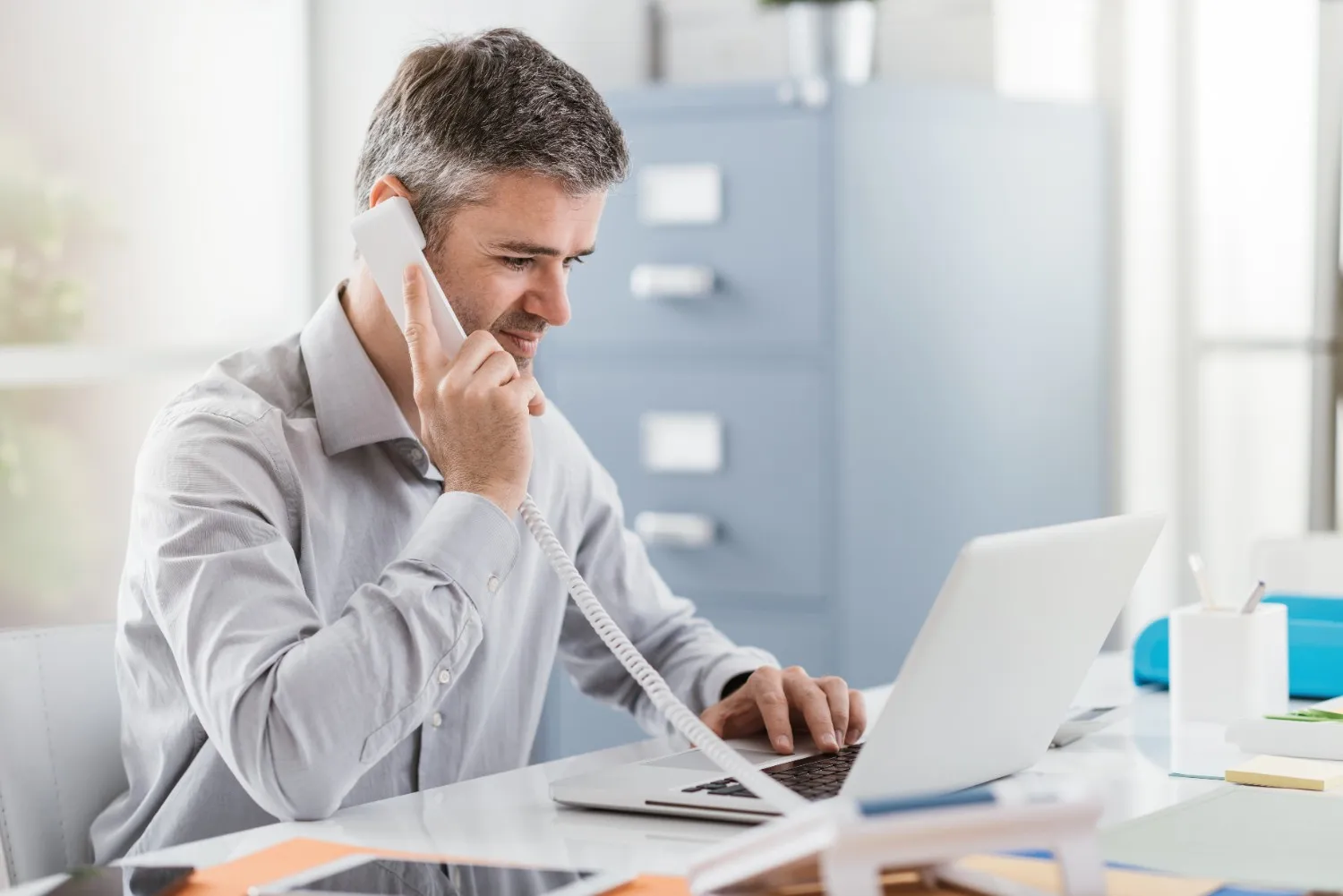 A male customer service representative talking on a landline phone while typing on a laptop.
