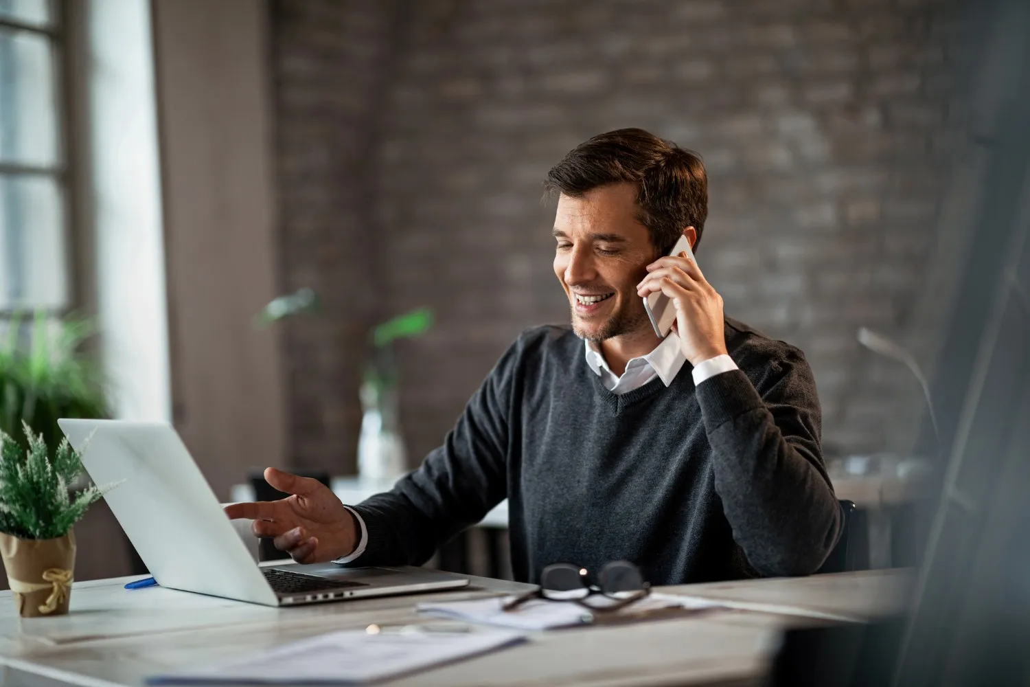 Smiling man working on a laptop while talking on a mobile phone at his desk.