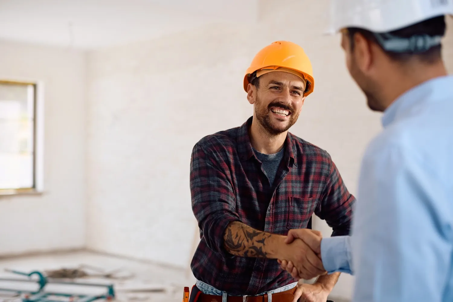  Two men are shaking hands in an unfinished interior room. One man, wearing an orange hard hat and a plaid flannel shirt with visible tattoos on his forearm, is smiling warmly while facing the camera. The other man is only visible from the shoulder back, wearing a white hard hat and a light blue shirt.