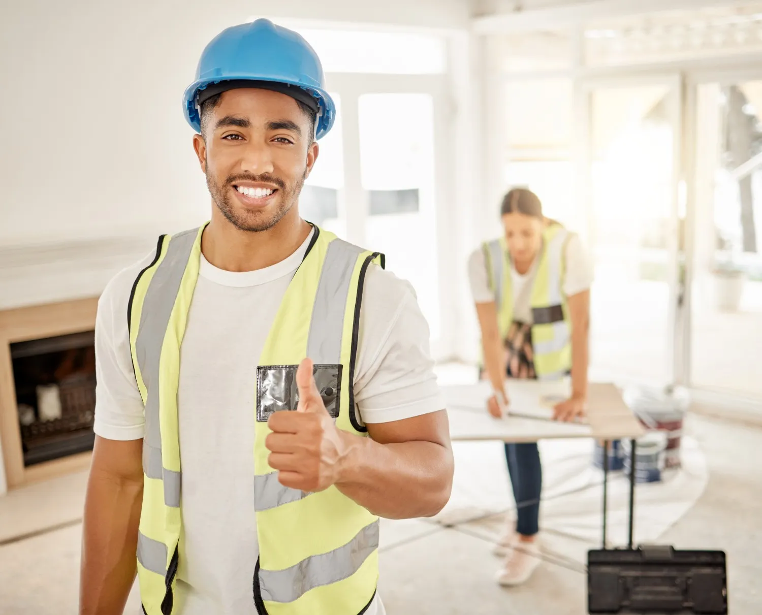  A smiling male construction worker in a blue hard hat and a high-visibility vest gives a thumbs up directly to the camera. He is standing inside a well-lit, unfinished home renovation project. In the blurred background, a female colleague in a vest is looking over blueprints on a table.
