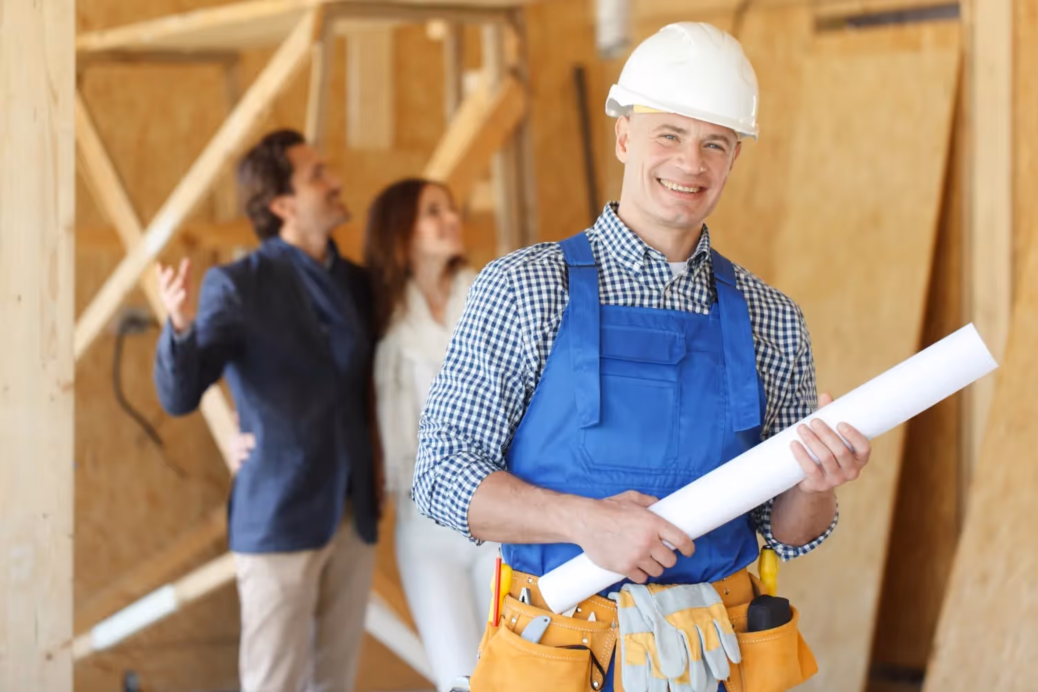  A smiling male construction contractor in a white hard hat, blue overalls, and a plaid shirt holds rolled-up blueprints. He is standing in a wooden-framed, unfinished interior. In the soft-focus background, a couple (a man in a suit jacket and a woman in a white shirt) look up and discuss the space.