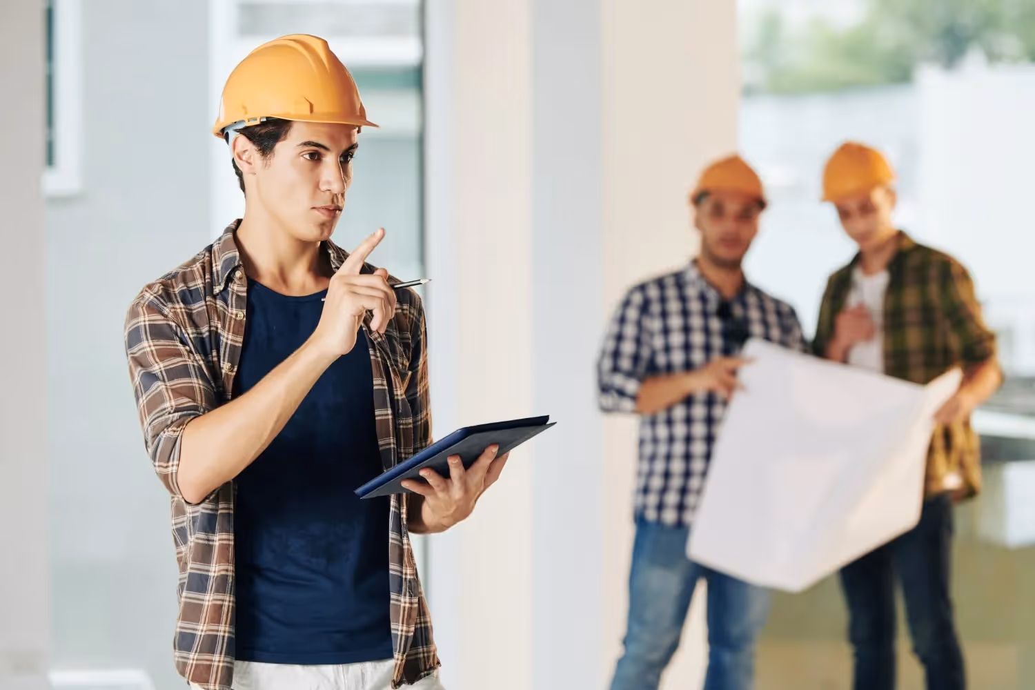 A young male construction foreman or architect in an orange hard hat and a plaid shirt stands in the foreground, holding a tablet and pointing his pen upwards, seemingly in thought. In the blurred background, two other men in orange hard hats review a set of blueprints.