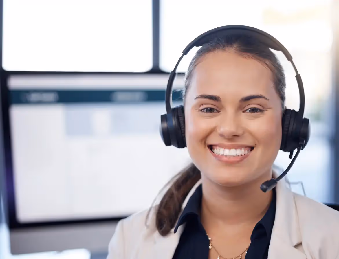 Close-up of a smiling female call center representative wearing a black headset in an office environment.