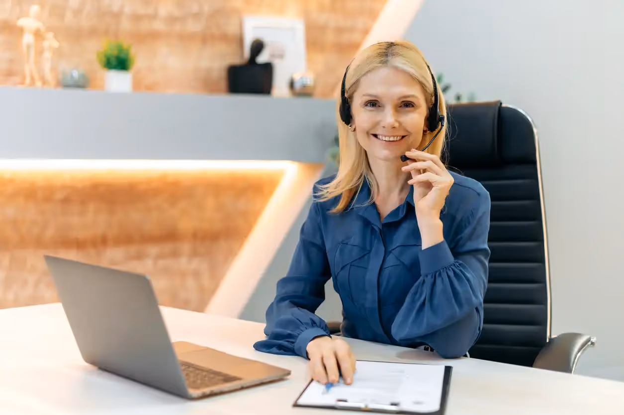 Professional blonde woman in a blue shirt and headset sitting at a desk with a laptop and paperwork.