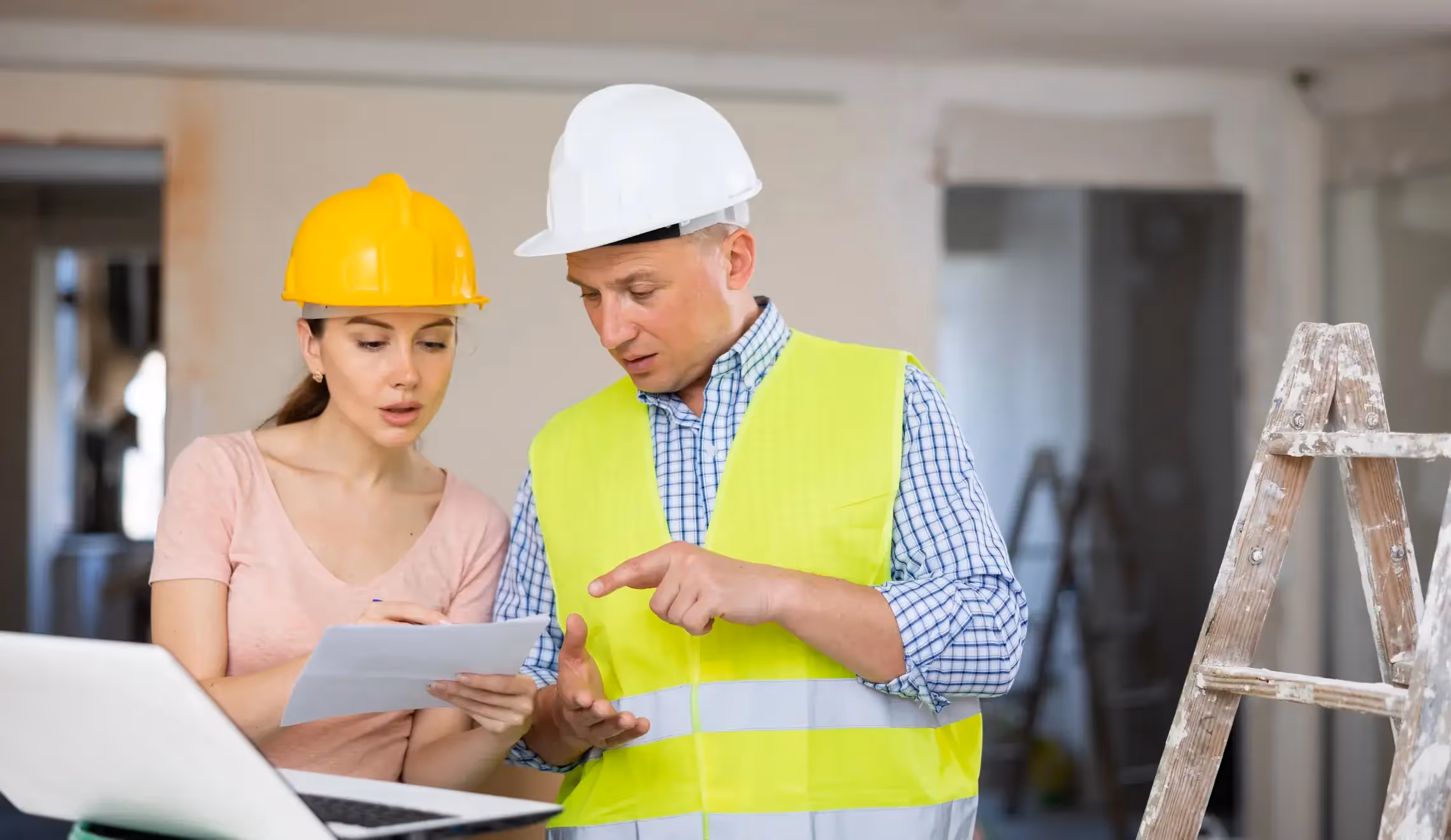 Contractor and client reviewing plans on a laptop at a construction site.