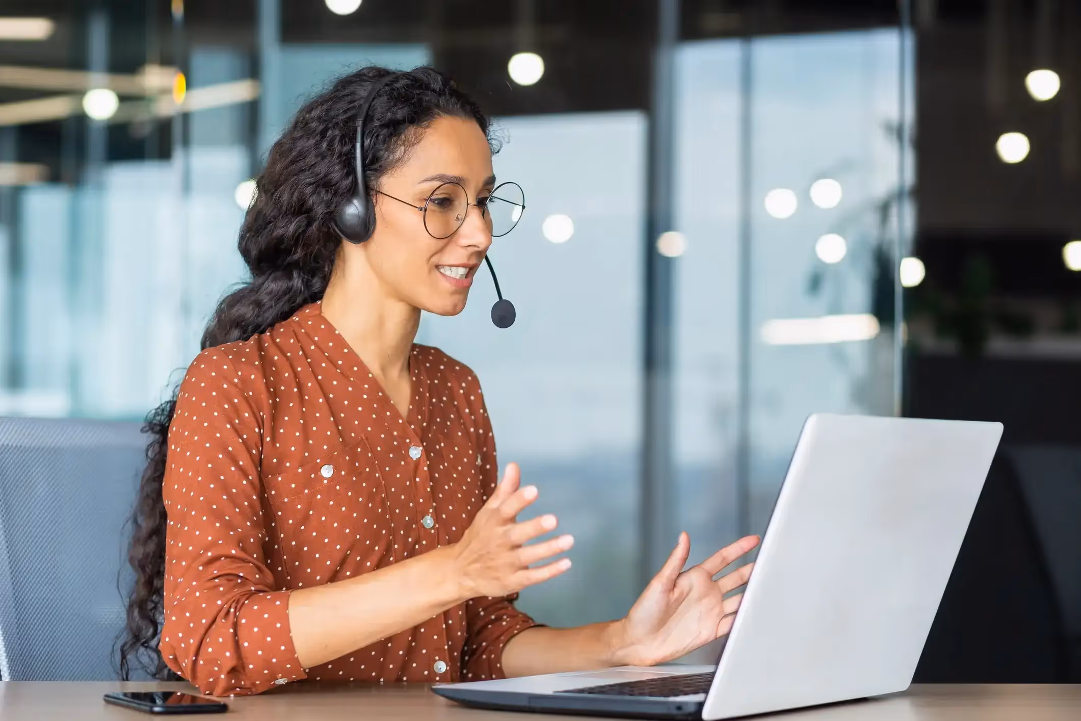 Happy female agent with curly hair and glasses in a video call on her laptop.