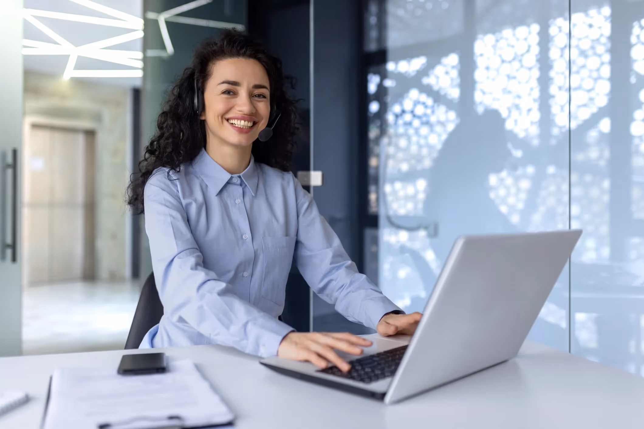 Cheerful woman in a headset smiling and typing on a laptop at her desk.