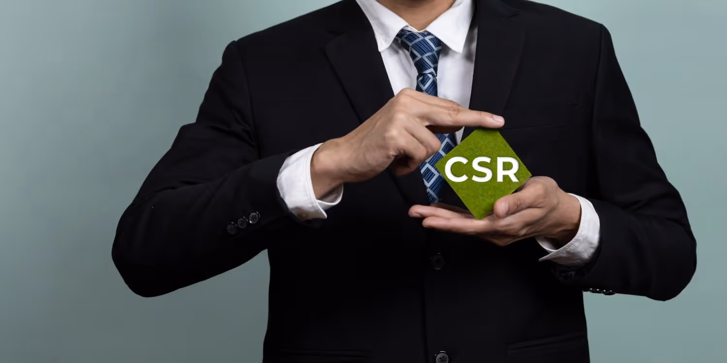 A man in a black business suit and patterned tie is holding a small, square, green card in his hands. The card, which has a texture resembling grass or moss, prominently displays the acronym CSR (Customer Service Representative or Corporate Social Responsibility) in white letters. The background is a solid light gray-blue.