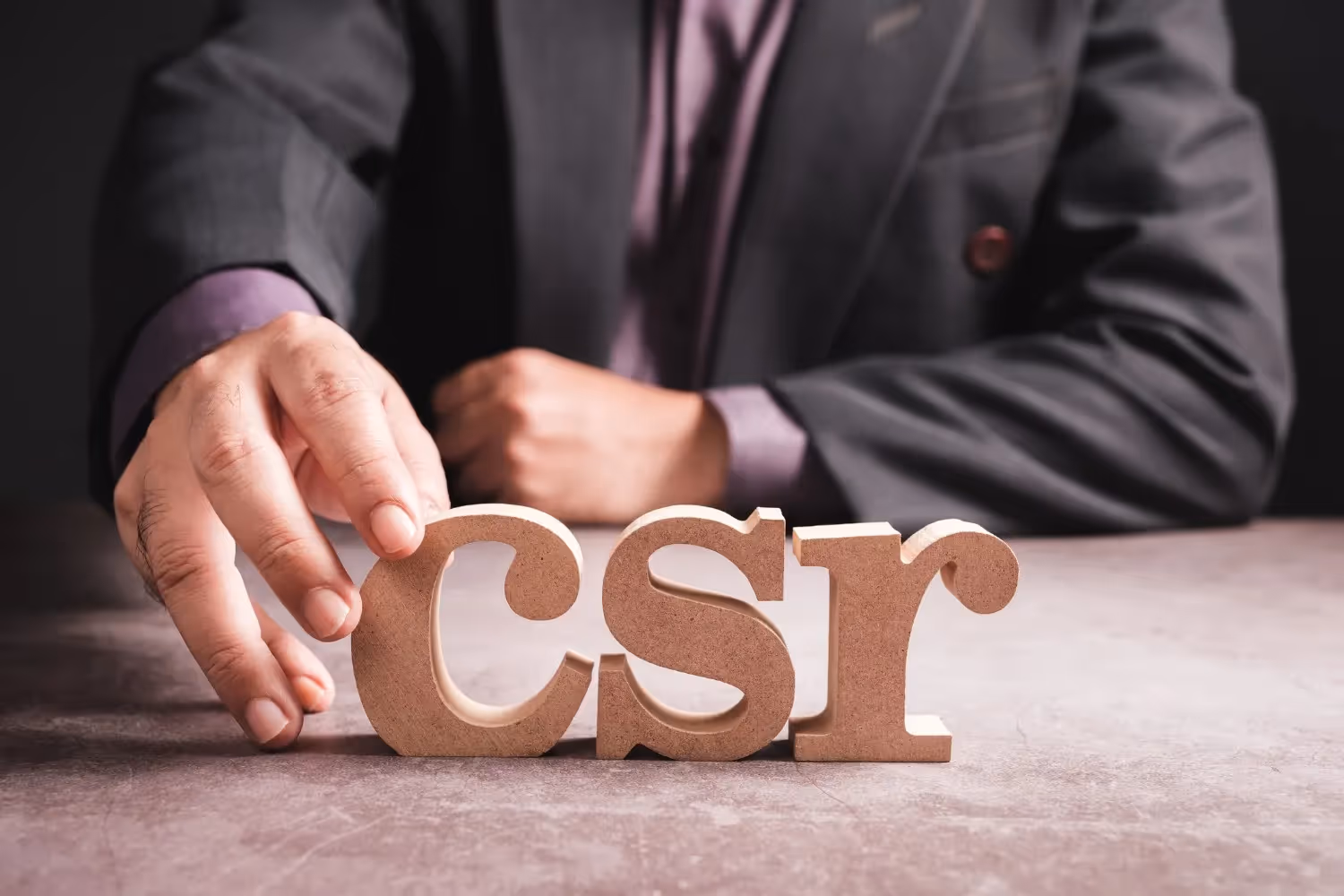  A person wearing a dark suit jacket and a purple shirt is seated at a table, reaching out to touch or adjust the first letter of the acronym "csr" spelled out in large, light brown wooden block letters on a textured dark surface. The image emphasizes the concept of CSR (Corporate Social Responsibility or Customer Service Representative).