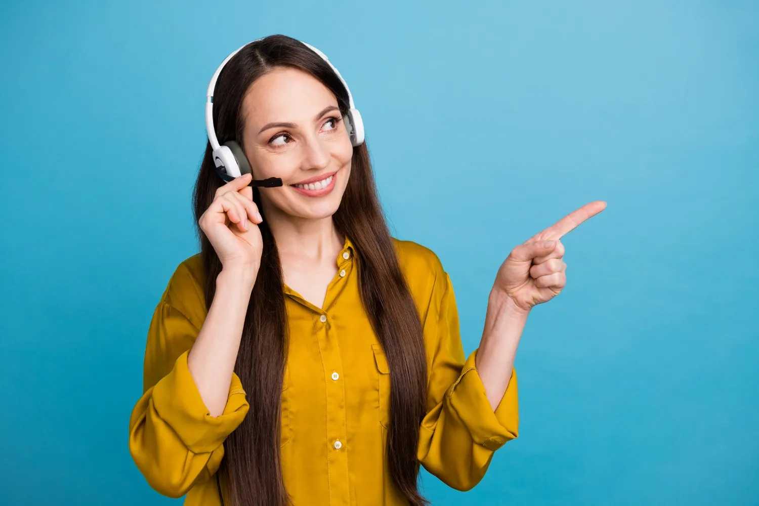  A cheerful woman with long, dark hair, wearing a mustard yellow shirt and white over-ear headphones with a microphone, is standing against a bright blue background. She is smiling, looking upwards, and pointing her right index finger to the upper right corner of the image, suggesting presentation or advertisement.