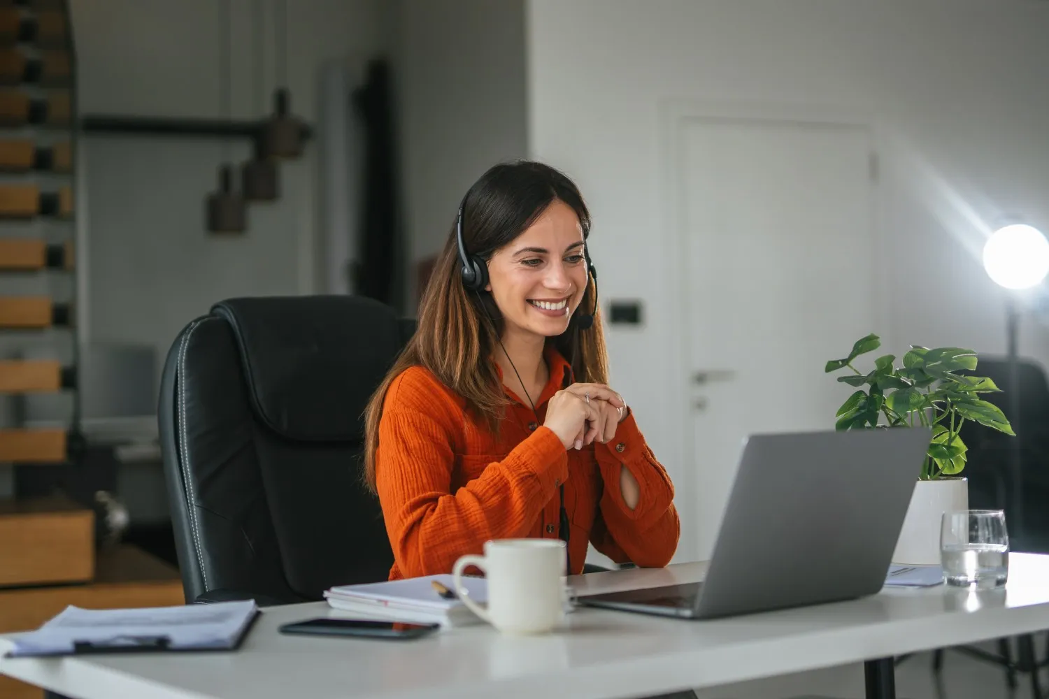  A smiling young woman with long dark hair is seated at a white desk in a home office, wearing a headset and an orange long-sleeved shirt. She is looking at her laptop screen, with her hands clasped, suggesting she is engaged in a video call or providing virtual customer support. A coffee mug, papers, and a potted plant are on the desk, with a staircase and a doorway visible in the background.