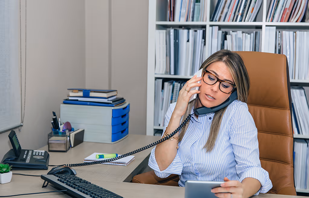 Woman in an office using a telephone and smartphone, managing customer calls for an answering service, showcasing multitasking in a business environment.
